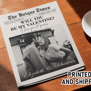 May include: A newspaper-style print with a black and white photo of a couple kissing. The headline reads "WILL YOU BE MY VALENTINE?" with the names "Elizabeth & Sebastian" below the photo. A cup of coffee sits on a saucer.