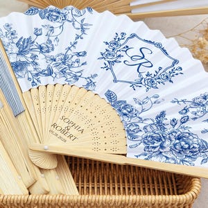 May include: White and blue floral printed fans with bamboo handles. One fan features the initials "S R" inside a floral frame. Another fan has the names "Sophia & Robert" and the date "05.28.2028" printed on it. The fans are in a woven basket.