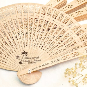 May include: A wooden hand fan with a delicate, intricate design. The fan is personalized with the text "We're married! Amelia & Michael 08.15.2026" and a palm tree graphic.