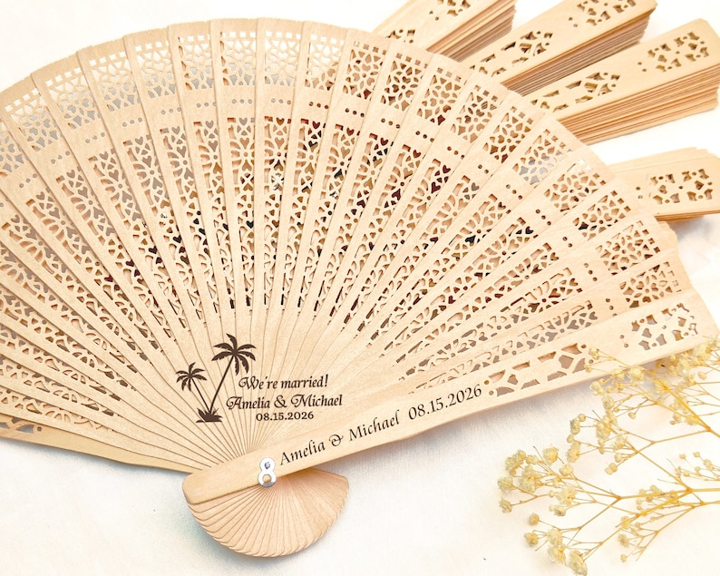 May include: A wooden hand fan with a delicate, intricate design. The fan is personalized with the text "We're married! Amelia & Michael 08.15.2026" and a palm tree graphic.