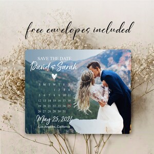 May include: Save the date card with a photo of a couple kissing in front of a mountain range. The card features a calendar with the date May 25, 2024 circled in red. The text "David & Sarah" is written in cursive with a heart symbol.
