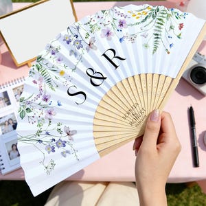 May include: A white folding fan with a floral design in purple, yellow, and green. The fan has the initials "S & R" and the names "Sophia Robert" printed on it. The fan is held by a person.