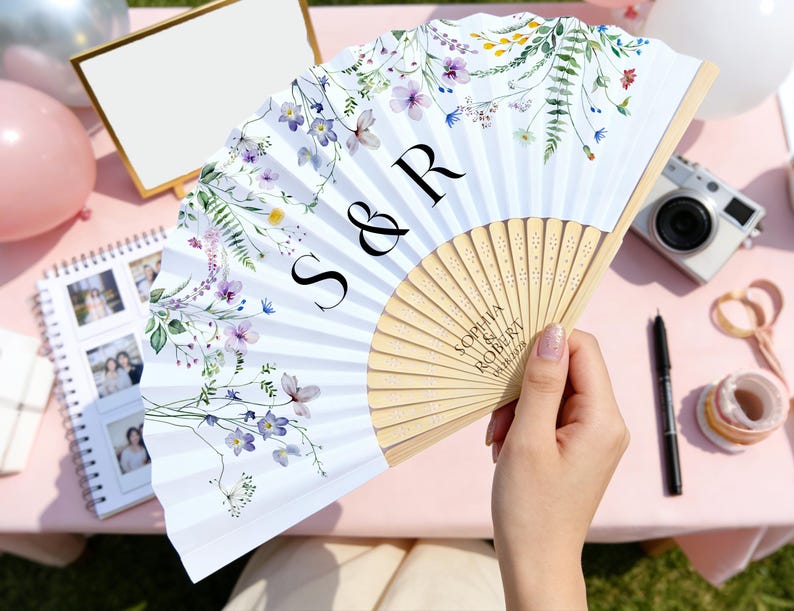 May include: A white folding fan with a floral design in purple, yellow, and green. The fan has the initials "S & R" and the names "Sophia Robert" printed on it. The fan is held by a person.