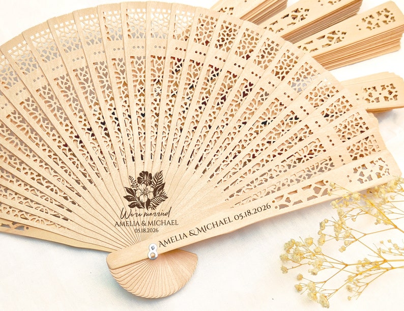 May include: A wooden hand fan with a floral design and the text "We're married Amelia & Michael 05.18.2026" engraved on the handle.