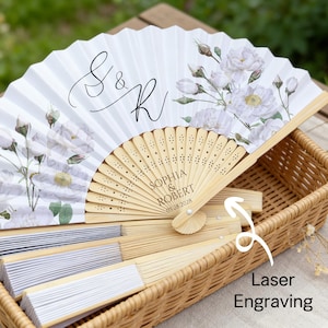 May include: White folding fan with floral design and laser engraving. The fan features the initials "S & R" and the names "Sophia & Robert" with a date. The fan is made of paper and wood and is displayed in a wicker basket.