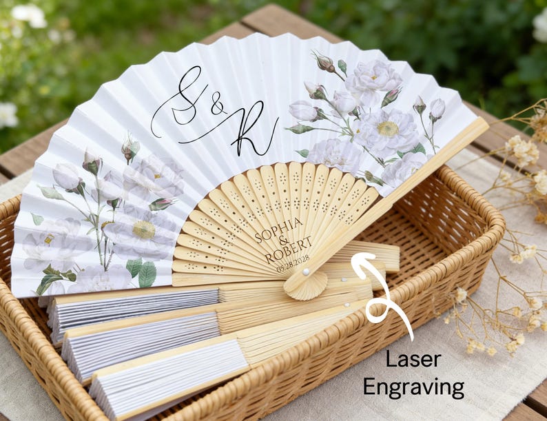 May include: White folding fan with floral design and laser engraving. The fan features the initials "S & R" and the names "Sophia & Robert" with a date. The fan is made of paper and wood and is displayed in a wicker basket.