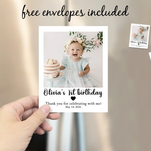 May include: A photo card held by a hand, featuring a smiling baby girl in a blue dress. The card reads "Olivia's 1st birthday" with a heart and the date "May 14, 2026." Two smaller cards are attached to a refrigerator. The text "free envelopes included" is above the card.