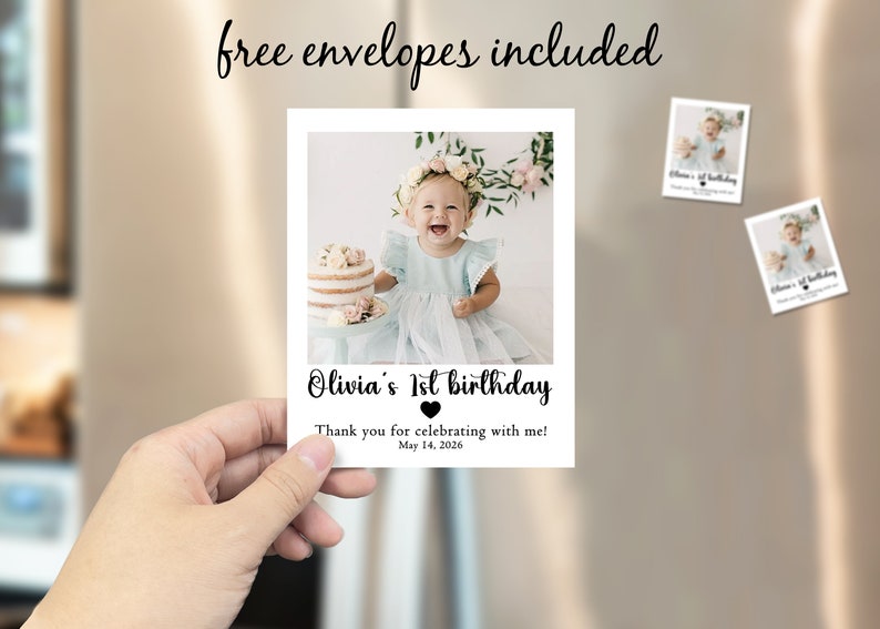 May include: A photo card held by a hand, featuring a smiling baby girl in a blue dress. The card reads "Olivia's 1st birthday" with a heart and the date "May 14, 2026." Two smaller cards are attached to a refrigerator. The text "free envelopes included" is above the card.