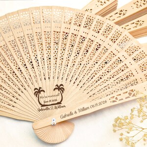 May include: A wooden hand fan with a laser engraved design of palm trees and the text "We're married! June 18, 2026 Gabriella & William" and "Gabriella & William 06.18.2026".