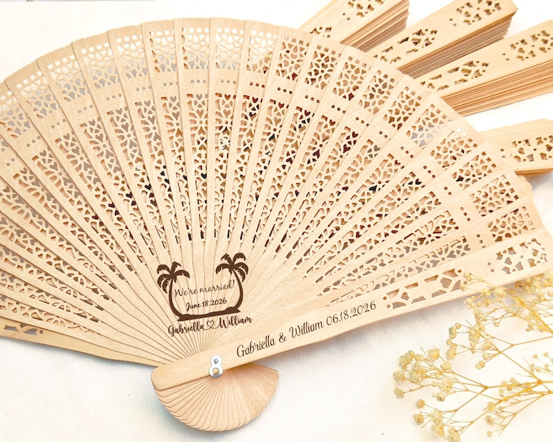 May include: A wooden hand fan with a laser engraved design of palm trees and the text "We're married! June 18, 2026 Gabriella & William" and "Gabriella & William 06.18.2026".