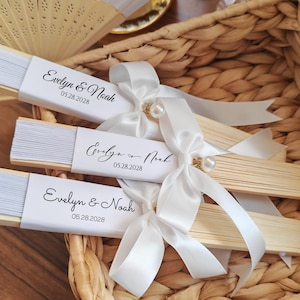 May include: Three wooden hand fans with white paper and the text "Evelyn & Noah 05.28.2028" are arranged in a woven basket. Each fan is adorned with a white satin ribbon and a pearl accent. The fans are a light beige color.