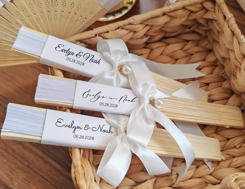 May include: Three wooden hand fans with white paper and the text "Evelyn & Noah 05.28.2028" are arranged in a woven basket. Each fan is adorned with a white satin ribbon and a pearl accent. The fans are a light beige color.