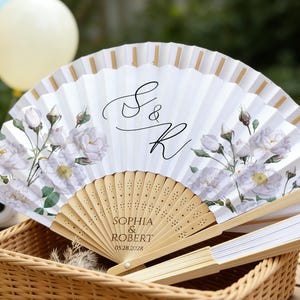 May include: White paper fan with floral design and gold accents. Features the names "Sophia & Robert" and the date "05.28.2028" printed on the wooden handle. The fan is displayed in a wicker basket.