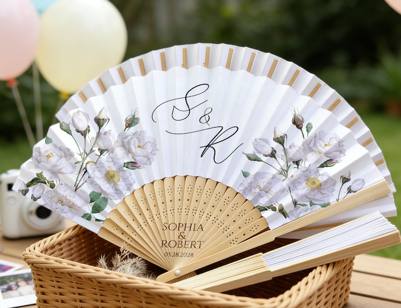 May include: White paper fan with floral design and gold accents. Features the names "Sophia & Robert" and the date "05.28.2028" printed on the wooden handle. The fan is displayed in a wicker basket.