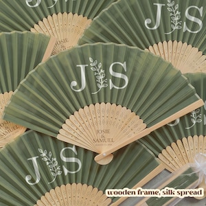 May include: A collection of sage green silk fans with wooden frames. Each fan features the initials "J S" in white, along with floral accents. Some fans display the names "Josie & Samuel" and a date. The fans are spread out.