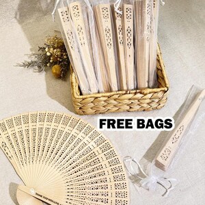 May include: A set of wooden folding fans with intricate cutouts, packaged in white organza bags. The fans are made of light brown wood and have a natural finish. The bags are tied with white ribbon. The text "FREE BAGS" is printed on a white label.