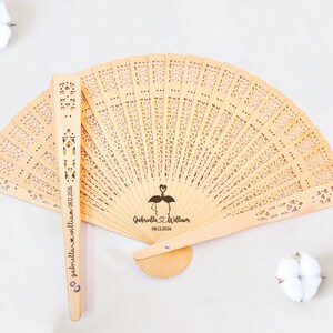 May include: A wooden hand fan with a delicate, intricate design. The fan is personalized with the names "Gabriella & William" and the date "08.12.2026" along with a flamingo illustration.