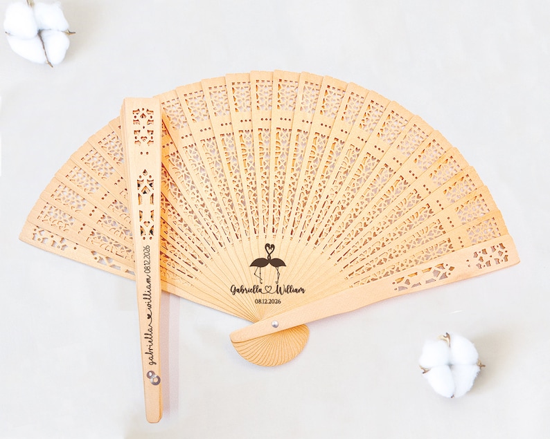 May include: A wooden hand fan with a delicate, intricate design. The fan is personalized with the names "Gabriella & William" and the date "08.12.2026" along with a flamingo illustration.