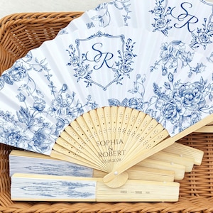 May include: White and blue floral printed folding fans with wooden handles. The fans feature a monogram and the names "Sophia & Robert" with the date 03.28.2028. The fans are displayed in a woven basket.