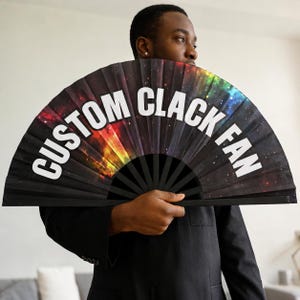 May include: A large handheld folding fan featuring a black background with a colorful nebula and star print. The text CUSTOM CLACK FAN is printed in bold white capital letters across the center of the fan, which has black ribs.