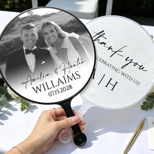 May include: Two round hand fans with black and clear plastic handles. One fan features a black and white wedding photo of a couple with the text Amelia & Hunter WILLAIMS 07.15.2028. The other fan displays the text thank you CELEBRATING WITH US! A | H.