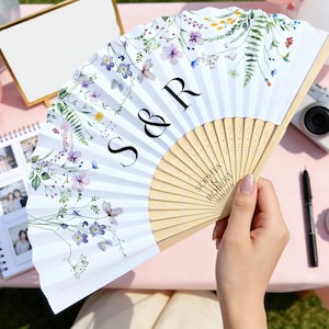 May include: A white folding fan with a floral design in purple, yellow, and green. The fan has the initials "S & R" and the names "Sophia Robert" printed on it. The fan is held by a person.