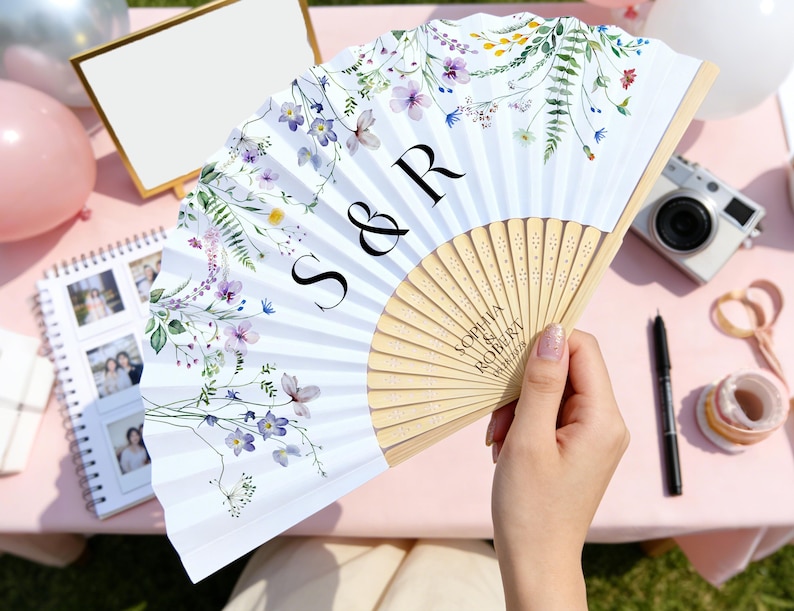 May include: A white folding fan with a floral design in purple, yellow, and green. The fan has the initials "S & R" and the names "Sophia Robert" printed on it. The fan is held by a person.