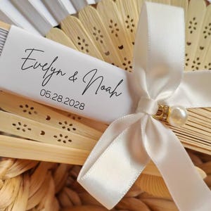 May include: A close-up of a decorative fan with a white ribbon and a pearl accent. The fan is made of light-colored wood and has a white paper label with the names "Evelyn & Noah" and the date "05.28.2028" printed on it. The fan is partially open.