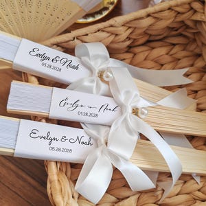 May include: Three wooden hand fans with white paper and the text "Evelyn & Noah 05.28.2028" are tied with white satin ribbons and pearl accents. The fans are in a woven basket, suggesting a wedding or event theme.