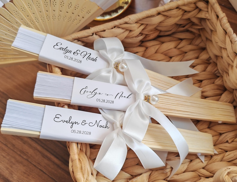 May include: Three wooden hand fans with white paper and the text "Evelyn & Noah 05.28.2028" are tied with white satin ribbons and pearl accents. The fans are in a woven basket, suggesting a wedding or event theme.