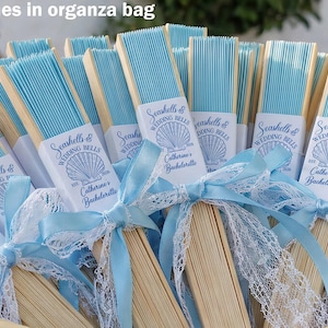 Könnte beinhalten: Eine Sammlung hellblauer Papierfächer mit Holzgriffen, gebunden mit hellblauen Bändern und weißer Spitze. Jeder Fächer hat ein weißes Etikett mit dem Text "Seashells & Wedding Bells" und "Catherine's Bachelorette".