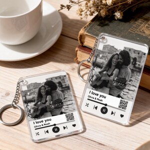May include: Clear acrylic keychains with a black and white photo of a couple playing guitar. The keychains have a music player interface design with the text "I love you Olivia & Noah". Each keychain has a silver chain and ring.