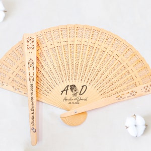 May include: A wooden hand fan with a delicate, intricate design. The fan is personalized with the initials "A & D" and the names "Amelia & Daniel" along with the date "08.15.2026".
