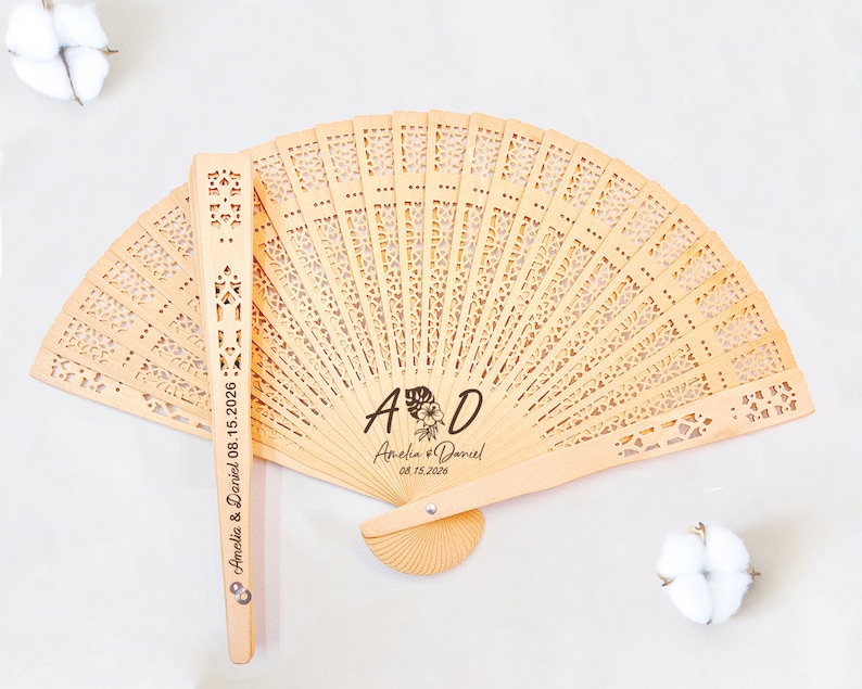 May include: A wooden hand fan with a delicate, intricate design. The fan is personalized with the initials "A & D" and the names "Amelia & Daniel" along with the date "08.15.2026".