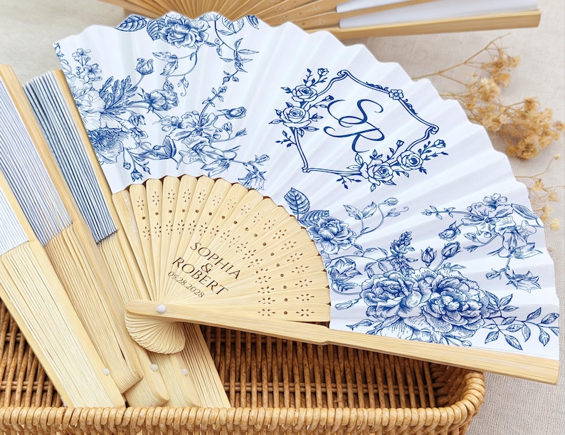May include: White and blue floral printed fans with bamboo handles. One fan features the initials "S R" inside a floral frame. Another fan is engraved with the names "Sophia & Robert" and the date "05.28.2028". The fans are displayed in a wicker basket.