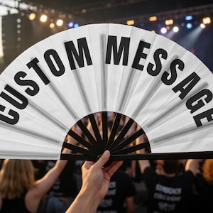 May include: A white handheld folding fan with black plastic ribs and the text CUSTOM MESSAGE printed in bold black capital letters across the fabric surface. The fan is fully extended in a semi-circular shape, displaying the clear text.
