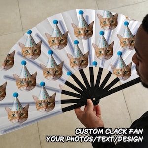 May include: A folding hand fan featuring a repeating pattern of orange cat faces wearing blue-tipped party hats against a white background. The fan has black plastic ribs and is held open to display the custom printed design and the text CUSTOM CLACK FAN YOUR PHOTOS/TEXT/DESIGN.