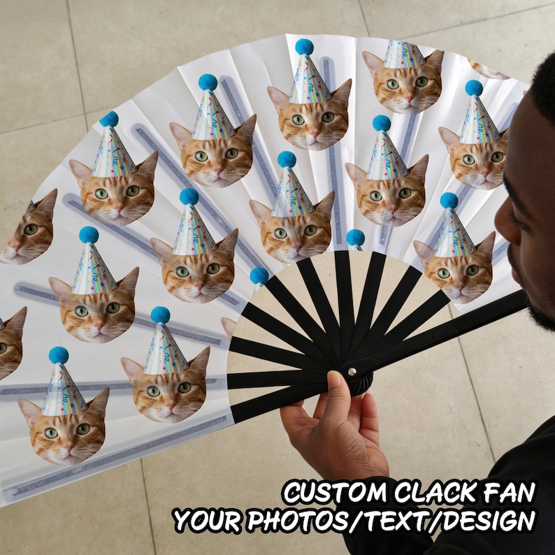 May include: A folding hand fan featuring a repeating pattern of orange cat faces wearing blue-tipped party hats against a white background. The fan has black plastic ribs and is held open to display the custom printed design and the text CUSTOM CLACK FAN YOUR PHOTOS/TEXT/DESIGN.
