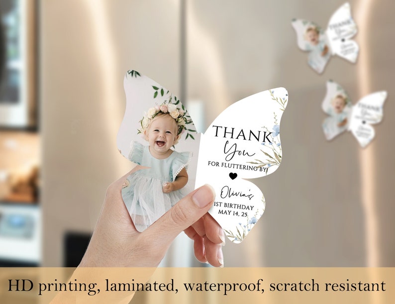 May include: Butterfly-shaped party favour with a photo of a baby girl wearing a floral crown. The favour reads "Thank You for Fluttering By" and "Olivia's 1st Birthday May 14, 25". The image also states "HD printing, laminated, waterproof, scratch resistant".