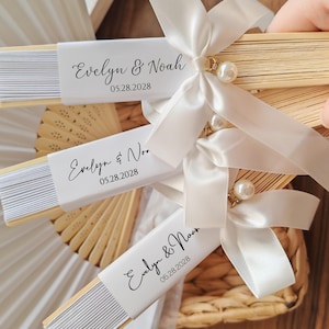 May include: Wooden folding fans with white paper inserts, personalized with the names "Evelyn & Noah" and the date "05.28.2028". Each fan is adorned with a white satin ribbon, a pearl, and a decorative gold accent. Ideal for wedding favors.