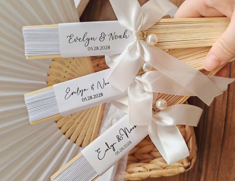 May include: Wooden folding fans with white paper inserts, personalized with the names "Evelyn & Noah" and the date "05.28.2028". Each fan is adorned with a white satin ribbon, a pearl, and a decorative gold accent. Ideal for wedding favors.