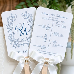 May include: Two white paddle-shaped wedding programmes with blue ink text and floral borders. The front features the monogram SM and The Smith. The back displays a timeline of events including marriage ceremony, dinner, and first dance with a white ribbon.