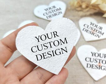 May include: A hand holding a white heart-shaped tag with the text "YOUR CUSTOM DESIGN" in black. Several other heart-shaped tags with the same text are visible in the background. The tags have a textured surface.