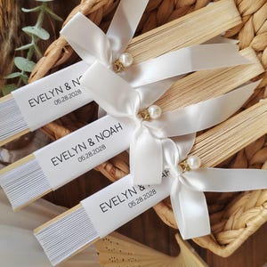 May include: Several personalized paper fans with white ribbon and pearl accents are arranged in a woven basket. Each fan has a white band with the names "EVELYN & NOAH" and the date "05.28.2028".