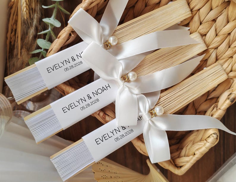 May include: Several personalized paper fans with white ribbon and pearl accents are arranged in a woven basket. Each fan has a white band with the names "EVELYN & NOAH" and the date "05.28.2028".