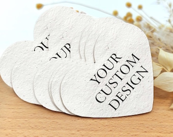 May include: A stack of heart-shaped paper coasters with the text "YOUR CUSTOM DESIGN" printed in black. The coasters have a textured, off-white appearance and are arranged on a wooden surface.