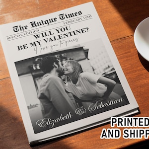 May include: A newspaper-style print with a black and white photo of a couple kissing. The headline reads "WILL YOU BE MY VALENTINE?" with the names "Elizabeth & Sebastian" below the photo. A cup of coffee sits nearby.