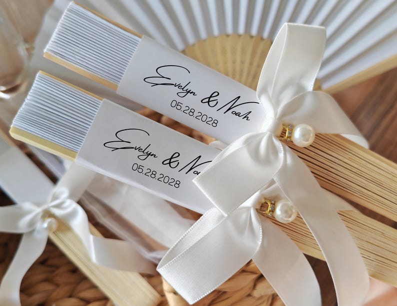 May include: Two personalized paper fans with white ribbon bows and pearl accents. Each fan has a white label with the names "Evelyn & Noah" and the date "05.28.2028" in black script. The fans are made of light-colored wood and white paper.