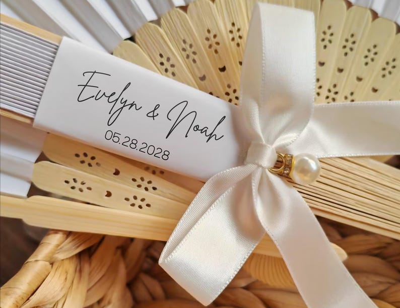 May include: A close-up of a decorative fan with a white ribbon and a pearl accent. The fan is made of light-colored wood and has a white paper label with the names "Evelyn & Noah" and the date "05.28.2028" printed on it. The fan is partially open.