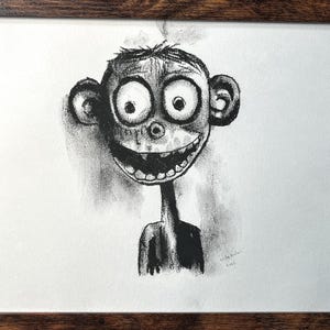 May include: A framed black and white drawing of a cartoonish figure with large eyes and a wide grin. The artwork is signed and dated, set within a dark brown wooden frame, suitable for wall decor.